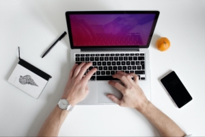 man typing on an Apple laptop with phone an orange and a pen surrounding the laptop on a white table.